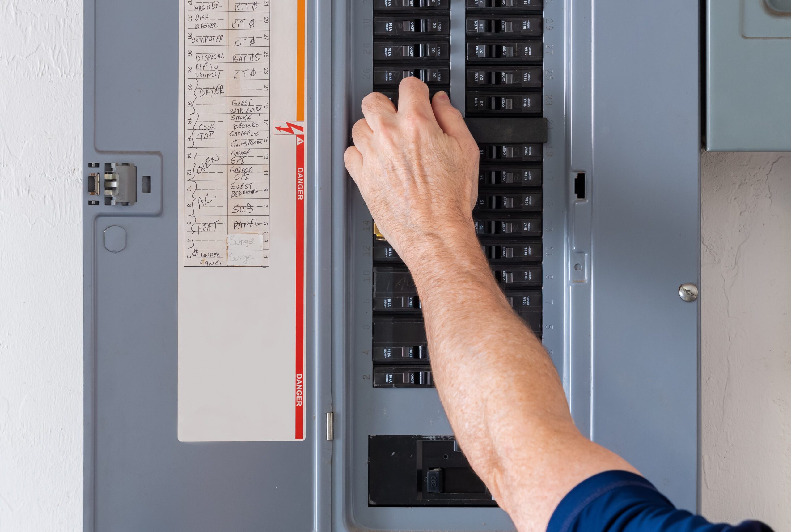 A person’s hand is flipping a switch inside an open electrical breaker panel with labeled circuits visible on the inside door.